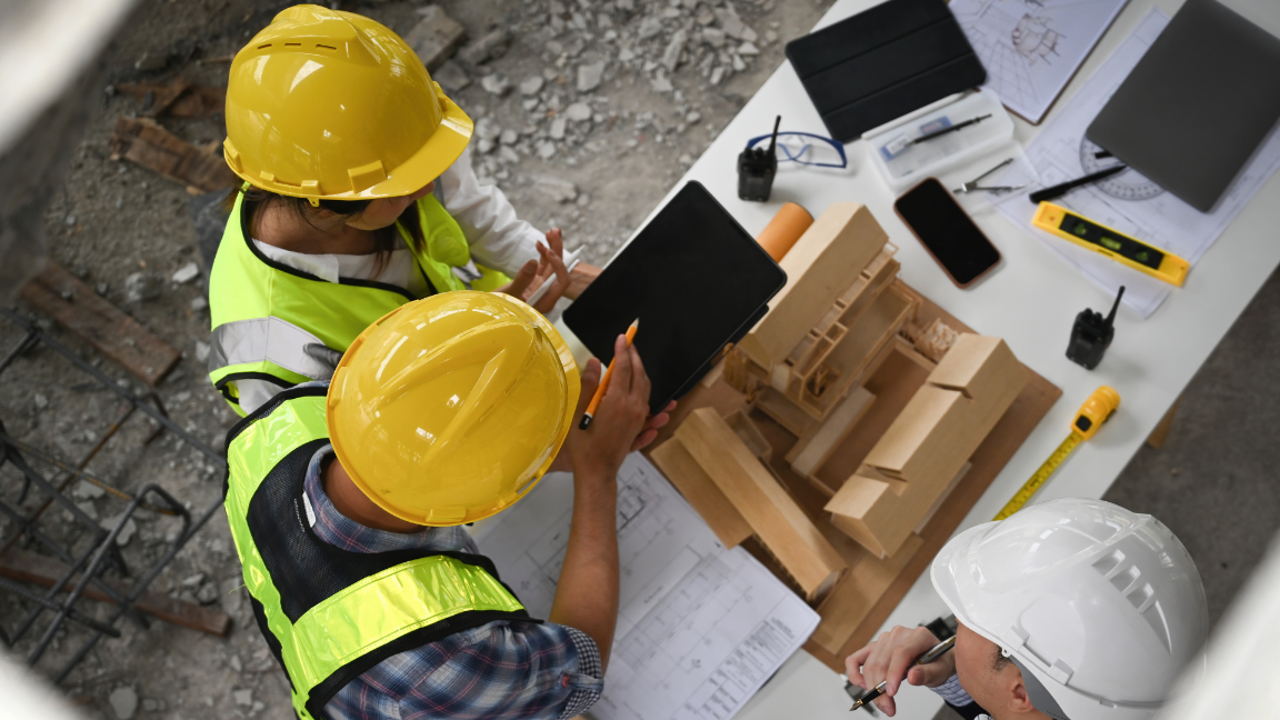 Three construction workers discussing a project. They have technical drawings, a model of the project, and IT equipment on the table.