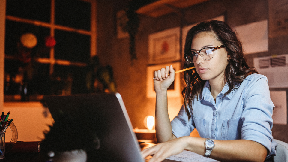 An image of a woman working at night. She is using a laptop, and reading a notebook on the table in front of her.