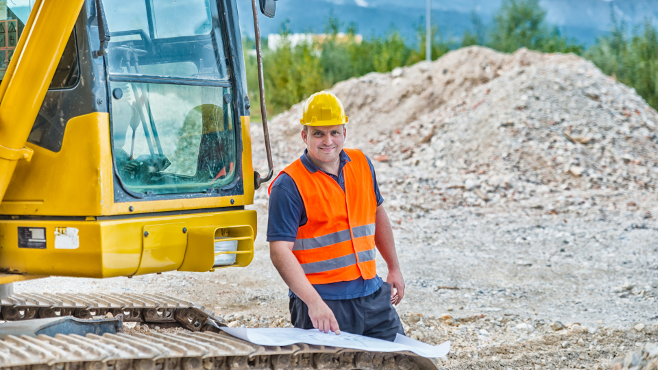 An image of a construction worker standing next to a 360 excavator machine on a construction site. He is wearing a hard hat and high-visibility vest, and is reading construction plans that are placed on the treads of the machine.