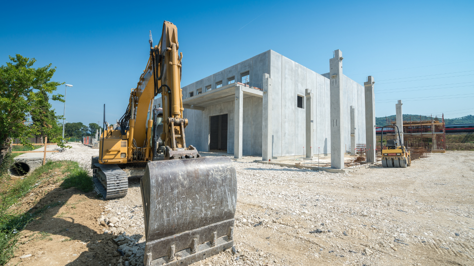 An image of a 360 excavator machine on a construction site. There is a concrete structure being built in the background behind the machine