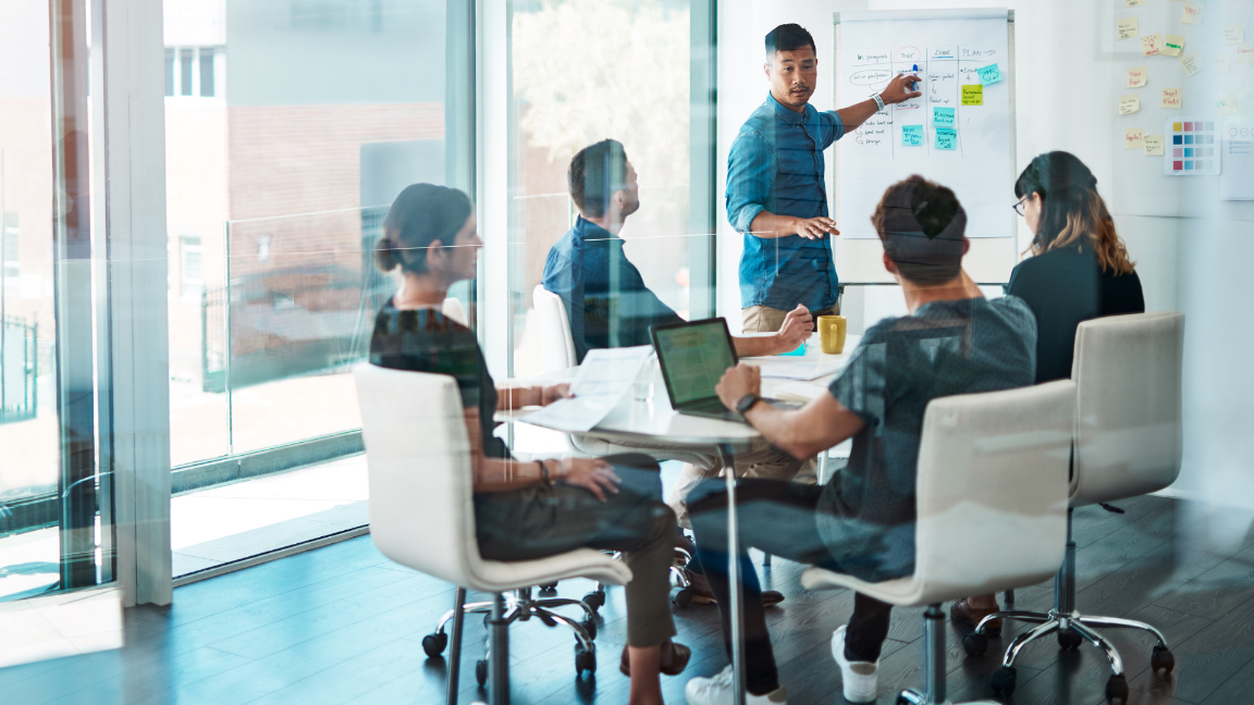 A business meeting taking place in a meeting room. Five people are sat around a table, and one person is stood up. The person standing is pointing a whiteboard and explaining the data.