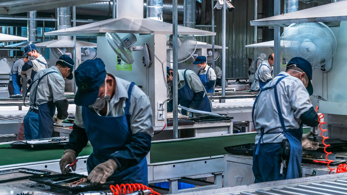 An image of multiple production workers in a factory. Each worker is at their own station, working on separate parts of the manufacturing proccess