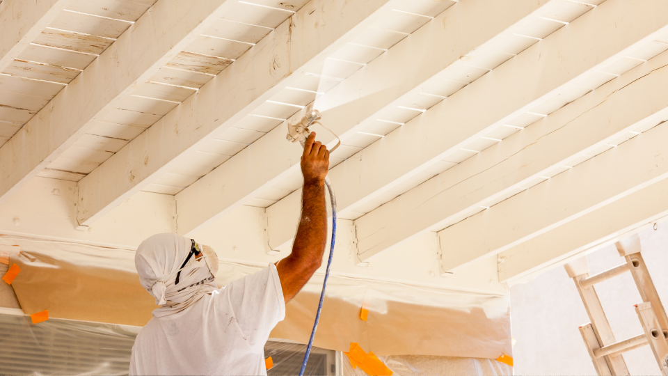 An image of a painter using a spray hose to paint a ceiling in a new building. He is wearing safety googles and a mask.