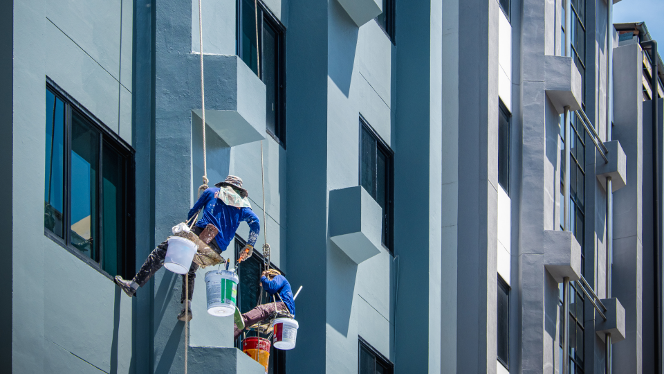 An image of two painters working on the outside of a new multi-story building. They are attached to safety lines and have their equipment on their specialist belts.