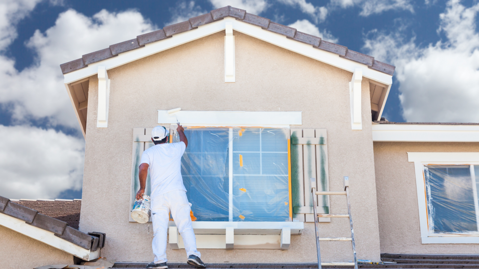 An image of a painter applying paint to the outside of a new house. He is using a roller to apply paint to the wooden frames of a window.