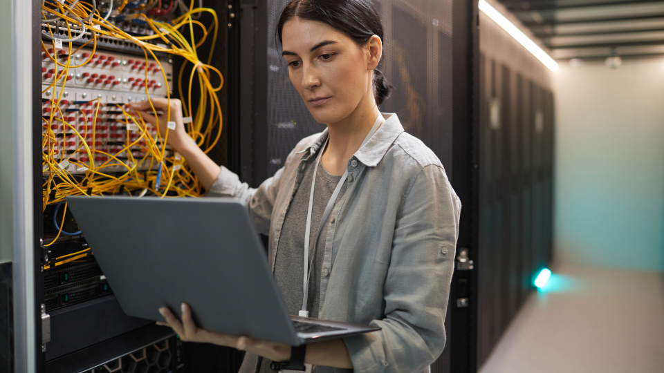 An image of a tech-support worker checking a server. They have a laptop plugged in to it and are reading outputs