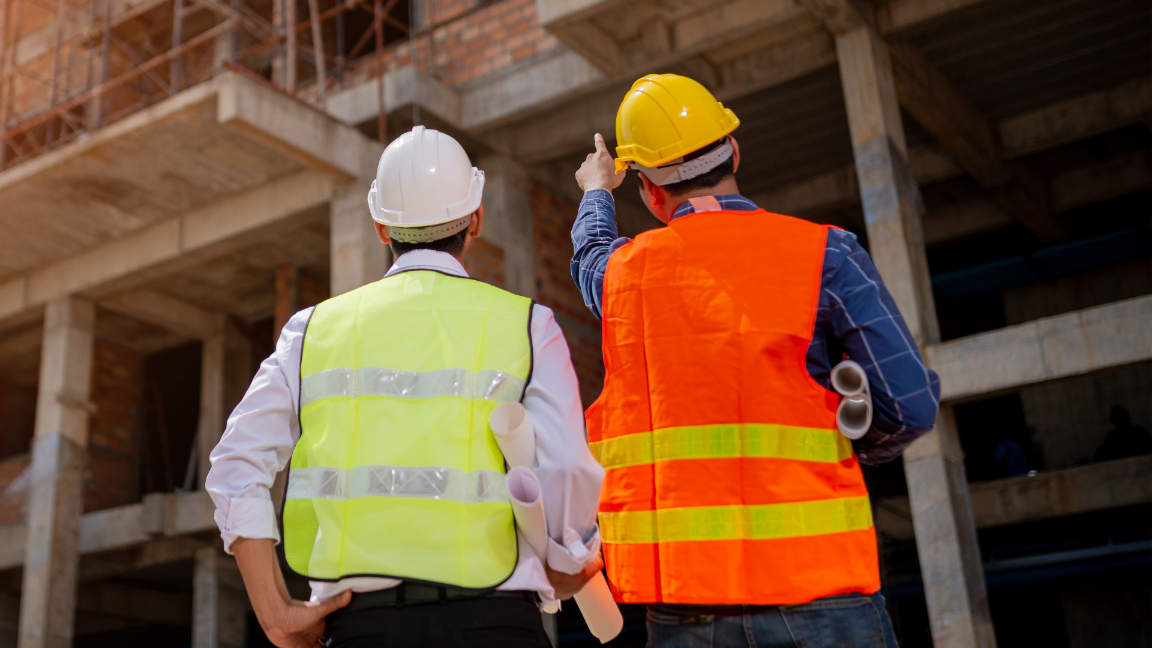 An image from behind of two construction workers pointing at at a construction site. They are wearing high-visibility jackets and hard-hats.