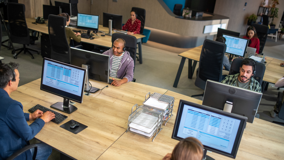 A high-angle image of several IT workers at multiple desks. They are on the phone to customers, and have information on their computer screens.