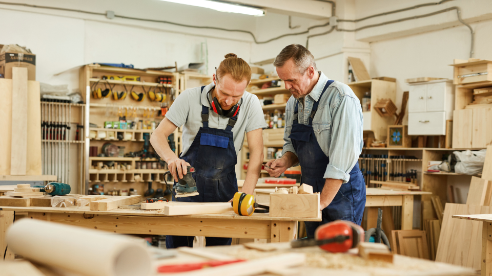 An image of two carpenters working on a piece of wood. One is sanding the surface of the wood, while the other is supervising. They are in a workshop and wearing blue overalls