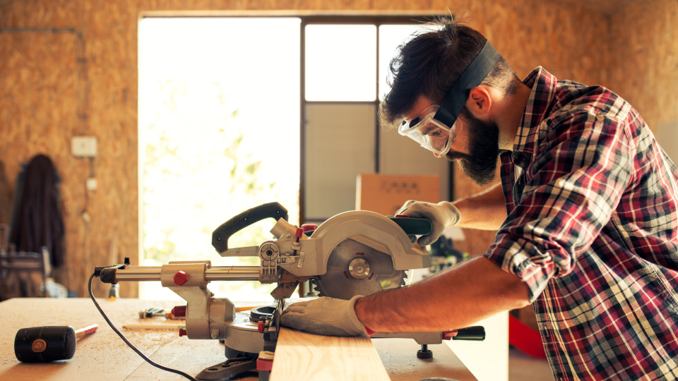 An image of a carpenter cutting a length of wood. He is wearing safety gloves and googles, and using a circular saw