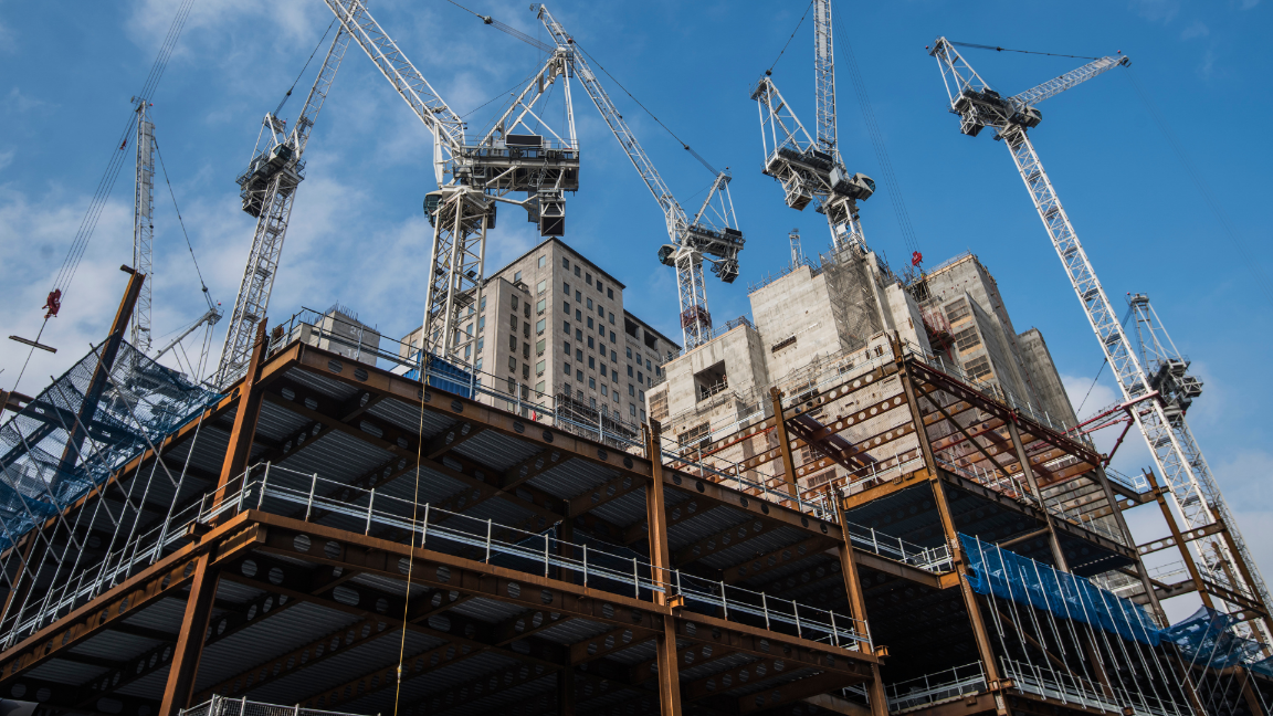 A low-angle image of a construction site. There is a concrete frame of a building, scaffolding, and cranes.