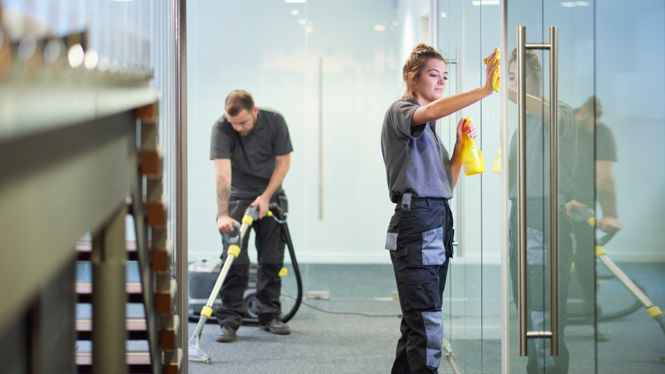 An image of two workers cleaning an office. A man is vacuuming the floor in the background, whilst a woman cleans a glass door in the foreground