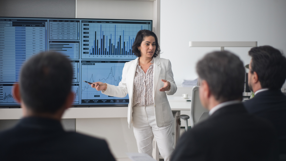 An image of a woman presenting financial data to a team. She is stood in front of a large display with data and charts on it.