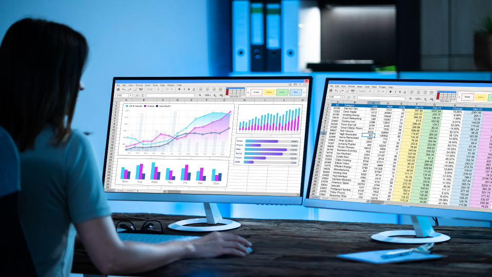 An image of a Financial Analyst looking at data on two computer screens. The screen on the left shows multiple charts, while the screen on the right has an excel sheet filled with data on it