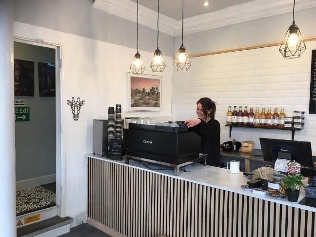 An image of a woman making coffee at a coffee machine. The machine is on the main counter of the coffee shop, and there are syrups on a shelf behind the woman