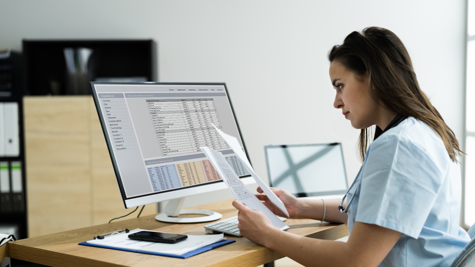 An image of a woman working on a computer. She is comparing data on pieces of paper to what is on her screen.