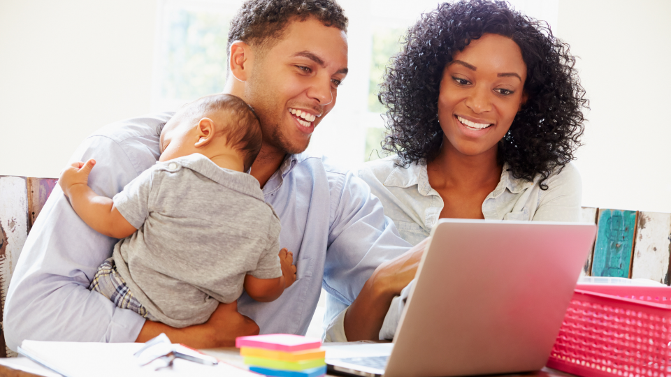 An image of a young family using a laptop. There is a woman and a man, and the man is holding a young baby