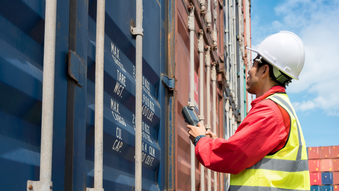 A worker checking a shipping container. He is wearing a hard-hat and high-vis vest. He is using a PDA to scan the container