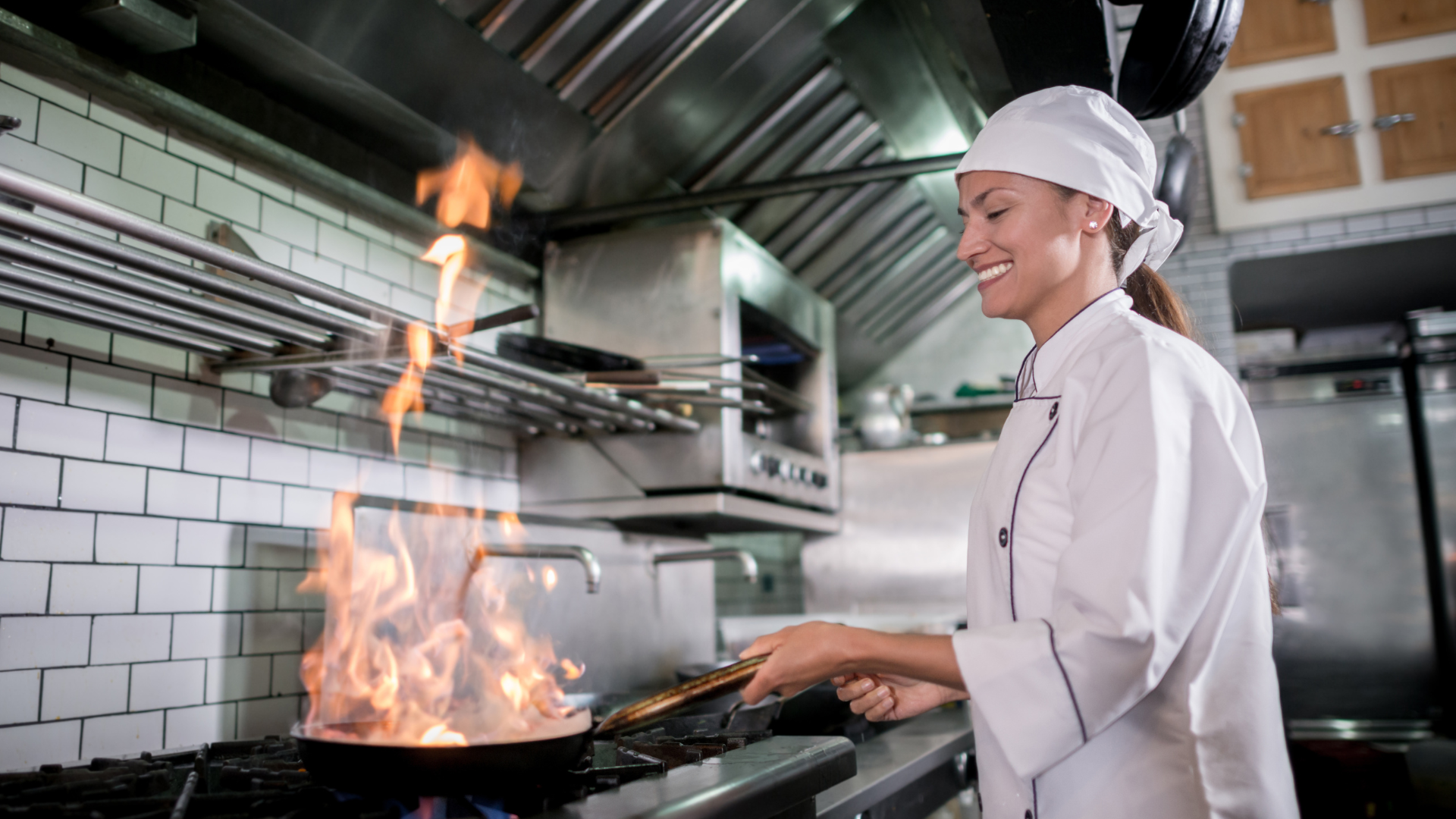 A female chef dressed in chef's whites and a hair protector. She is frying food in a restaurant kitchen and has a smile on her face