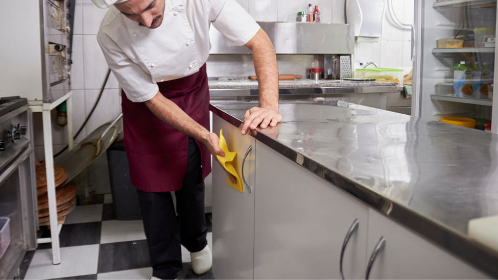 A kitchen porter wiping storage cupboards in a restaurant kitchen