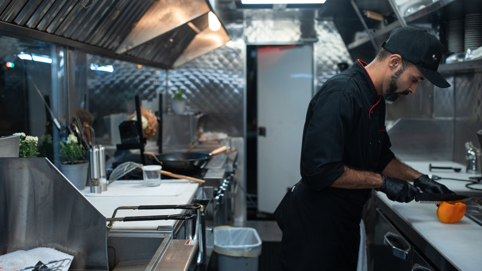 A lone chef chopping orange peppers in a restaurant kitchen. He is wearing gloves and a hat