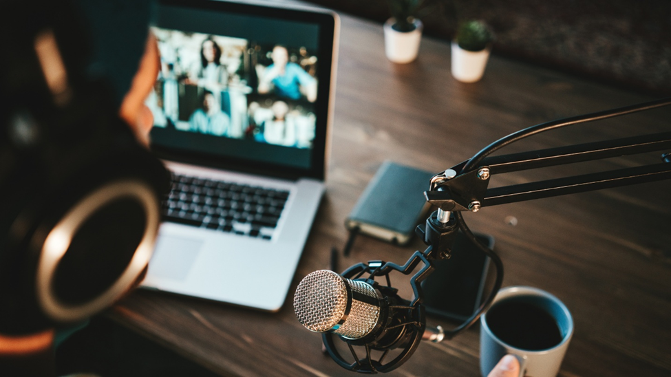 A person recording audio at their desk with a mounted microphone. There is a coffee cup, notepad, and laptop on the desk.