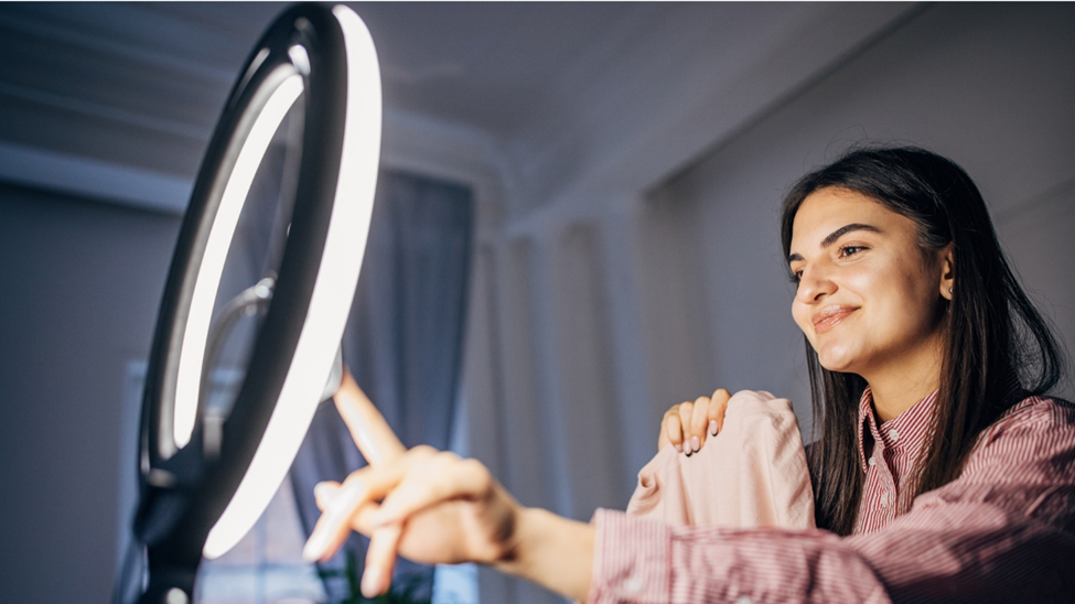 A woman recording a video on a smartphone with a ring light. The woman is holding up a product to show to the camera