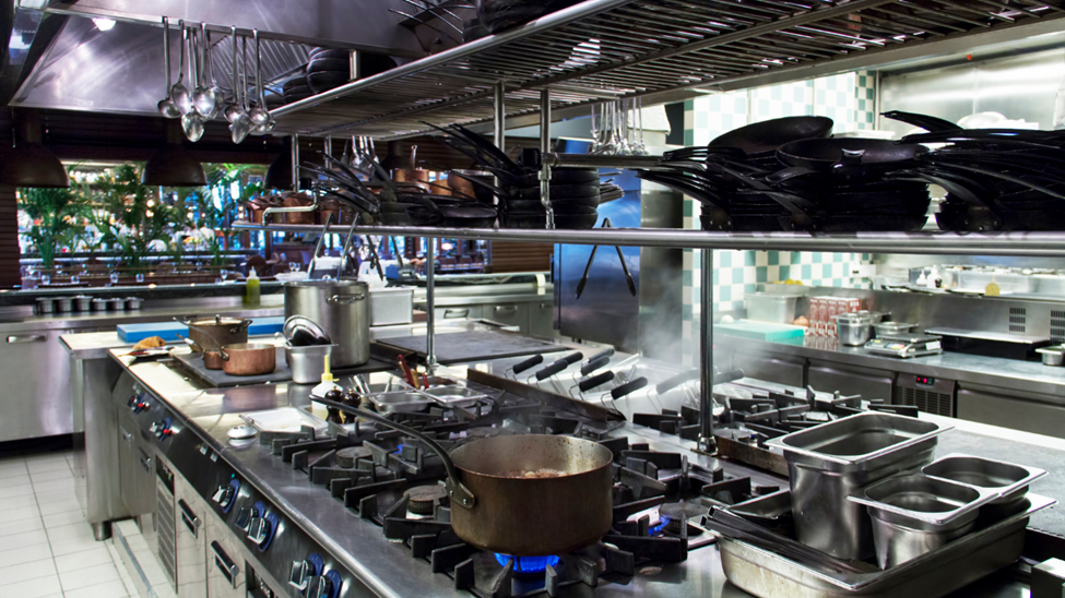 A wide-view of a commercial kitchen, with food cooking on a stove. Pots and pans and other kitchen equipment is stored around the kitchen