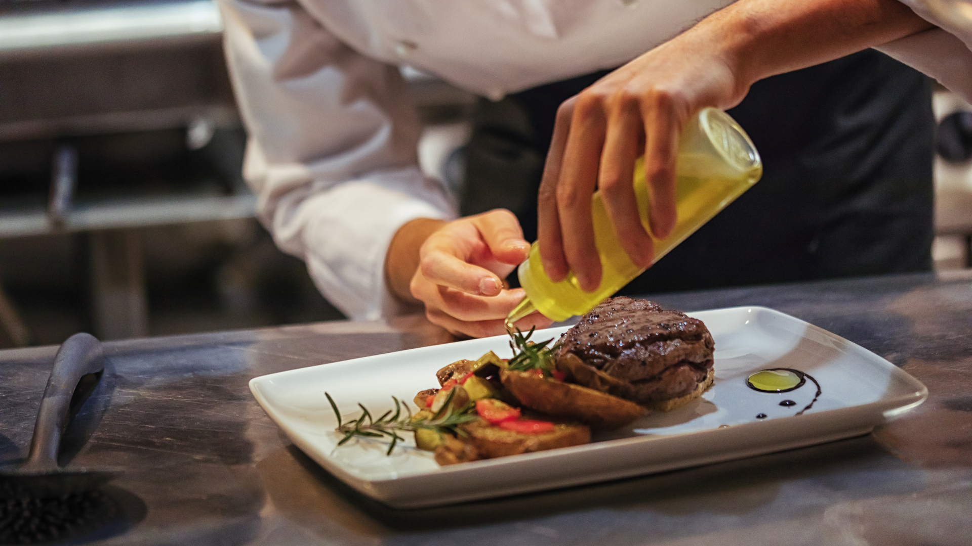 A chef putting the finishing touches on a burger, drizzling some oil over the potatoes on the plate