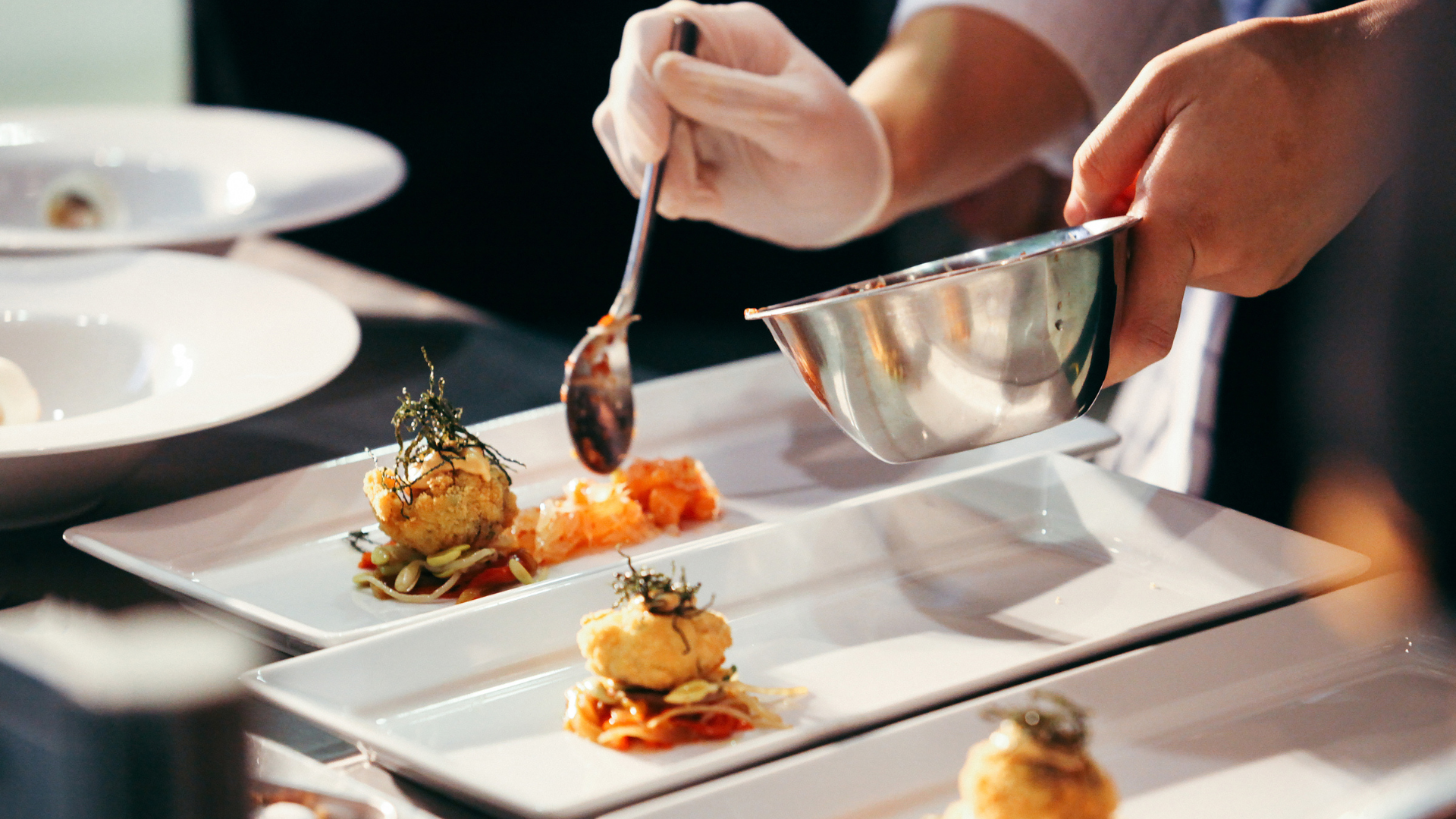 A chef adding components of a dish to several plates. They have a silver bowl with the food in it, and are putting it on the plates with a spoon.