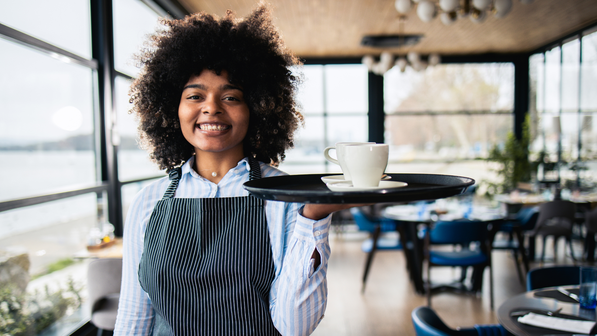 A waitress holding a tray with two coffee cups on it. She is wearing a striped apron and a blue shirt