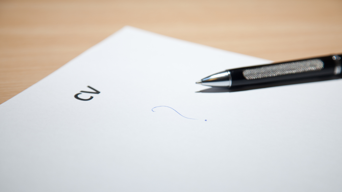 A sheet of paper on a wooden desk with a pen on it. The paper has the word "CV" on the top of the page, and nothing else except for a question mark in the middle of the page.