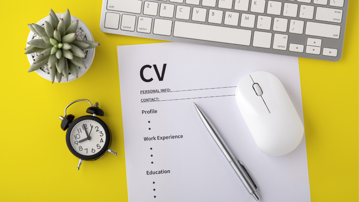 A CV on a yellow desk, with a pen, mouse, and keyboard on it. The CV has headings on it, but no details under the headings.