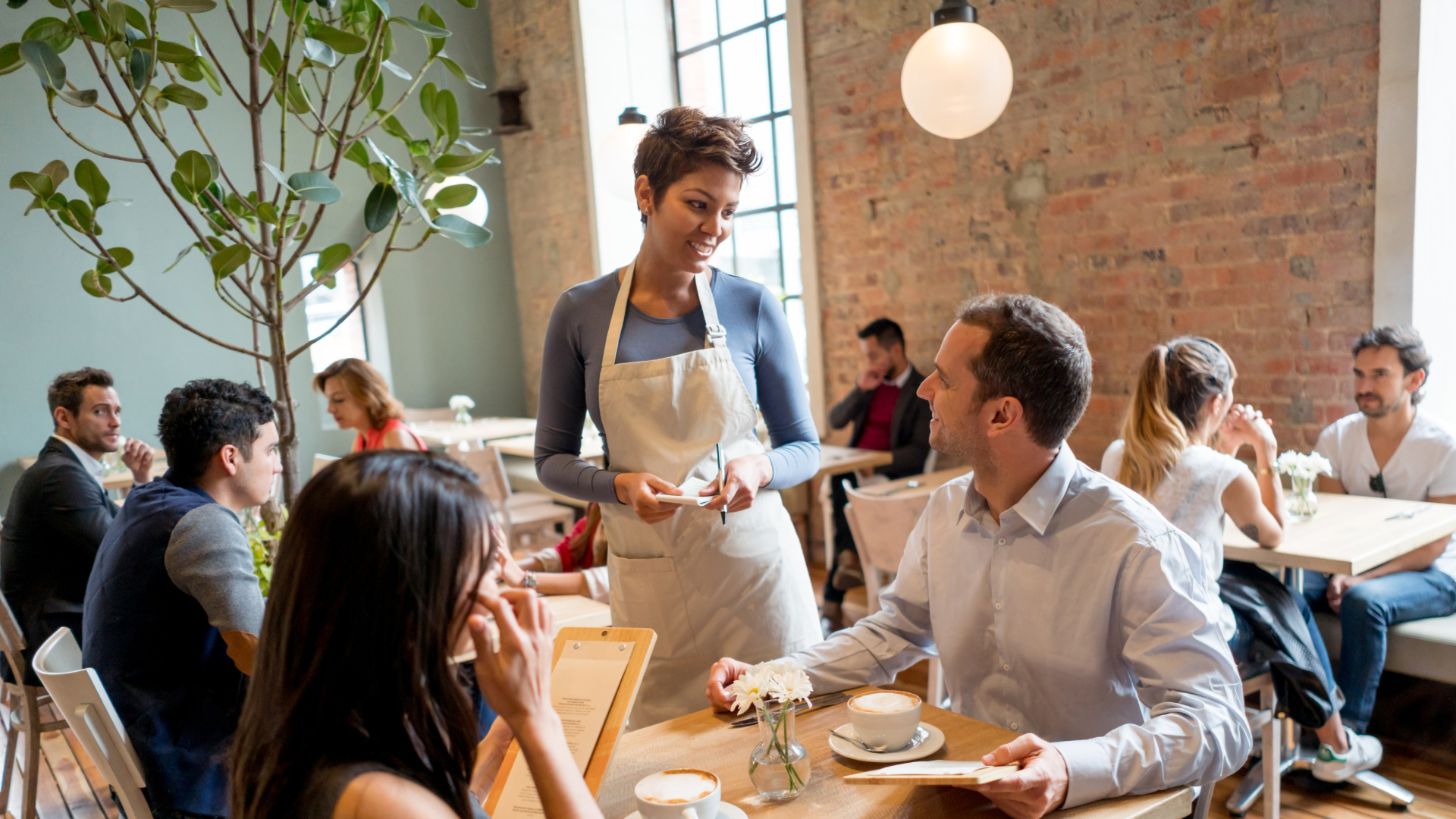 A waitress taking a customer's order at a restaurant. The waitress is holding a pen and a notepad. There are other customers sat at tables in the background