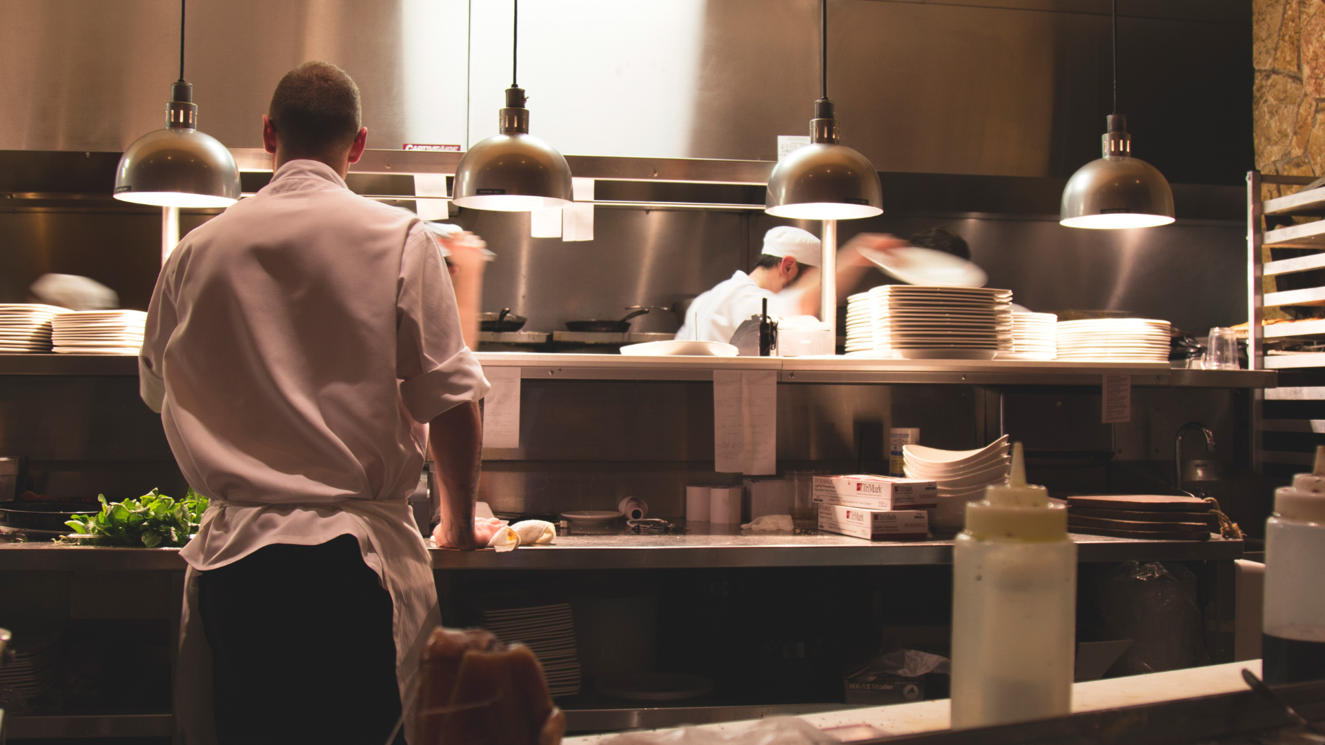 A chef by himself in a restaurant kitchen. It is dimly lit and people are rushing around him