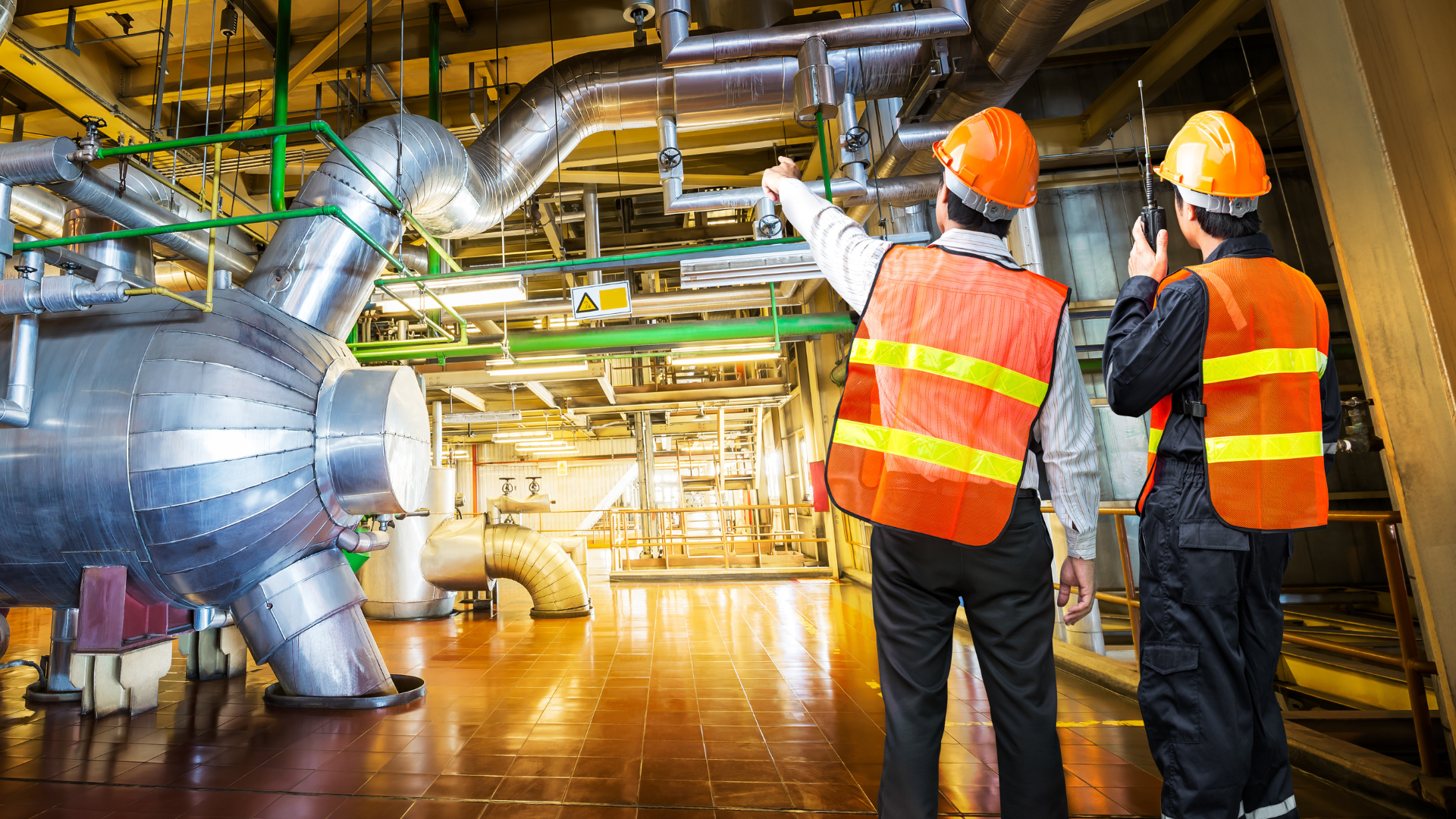 Two workers inspecting a green energy plant. They are wearing hard hats and high-vis jackets. The plant has large silver machines and green pipes running along the ceiling