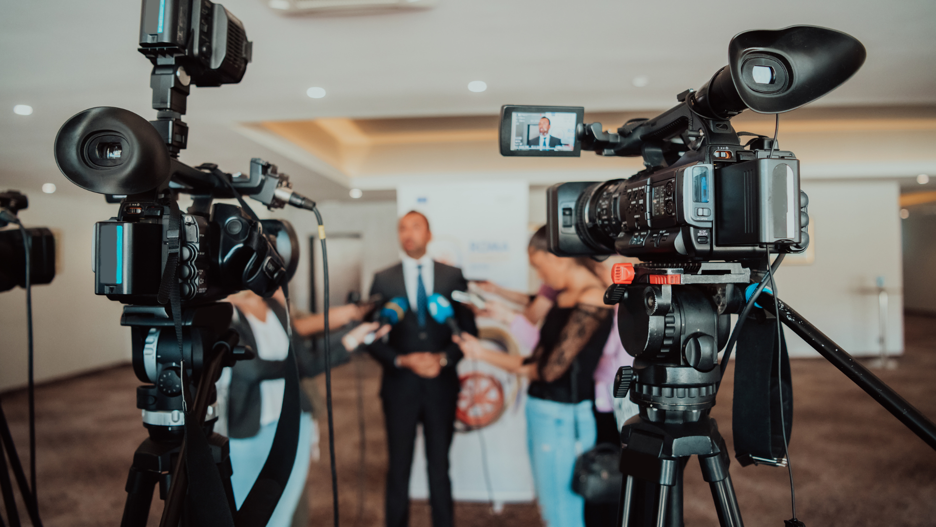 A man giving a press conference. He is stood in front of a sign, and multiple journalists are holding microphones up to him. Two large cameras are in the foreground, recording the event.