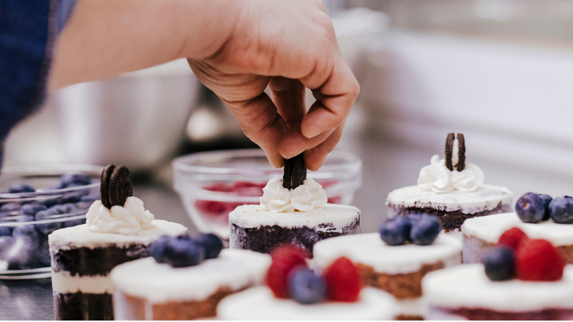 A range of small cakes on a commercial kitchen counter with various toppings