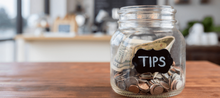 A glass jar on a wooden table. The jar has the word "Tips" written on it. There are coins and banknotes in the jar.