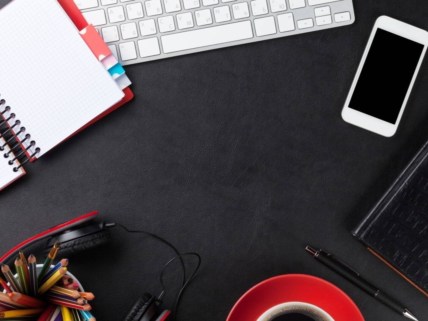 A notebook, keyboard, smart phone, and headset on a black desk