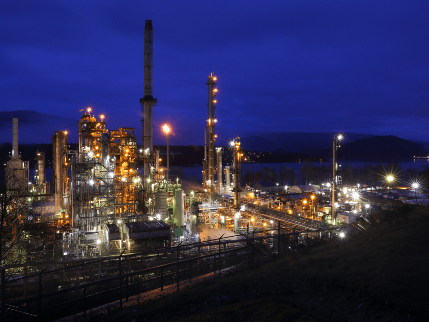 An image of a factory at night. The factory has many lights on the various chimneys.