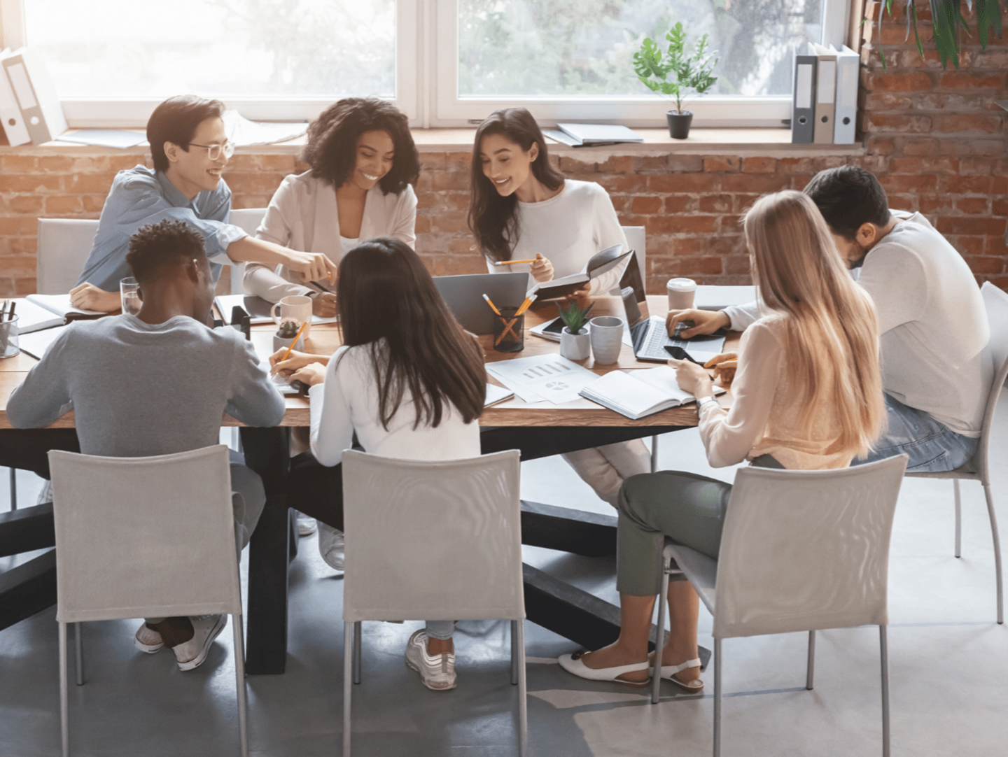 A diverse group of people working on a marketing project. They are sat at a wooden table that has laptops and papers on it.