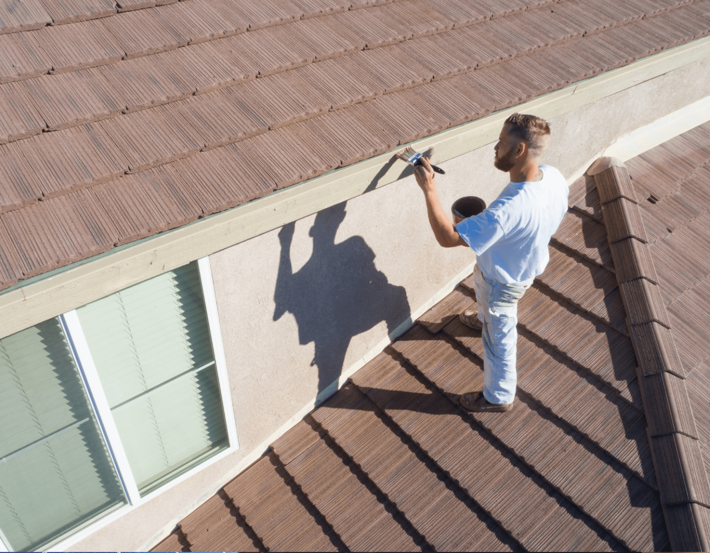 A high-angle image of a painter applying paint to the outside of a new house. He is stood on the roof and painting a wooden beam.