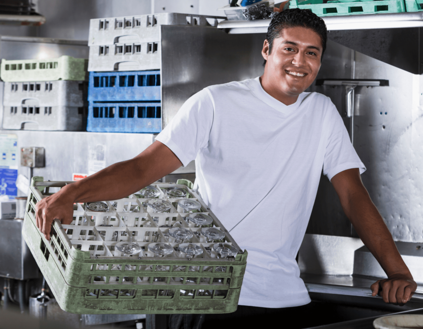 A kitchen porter holding a crate of glasses, standing near a sink in a kitchen