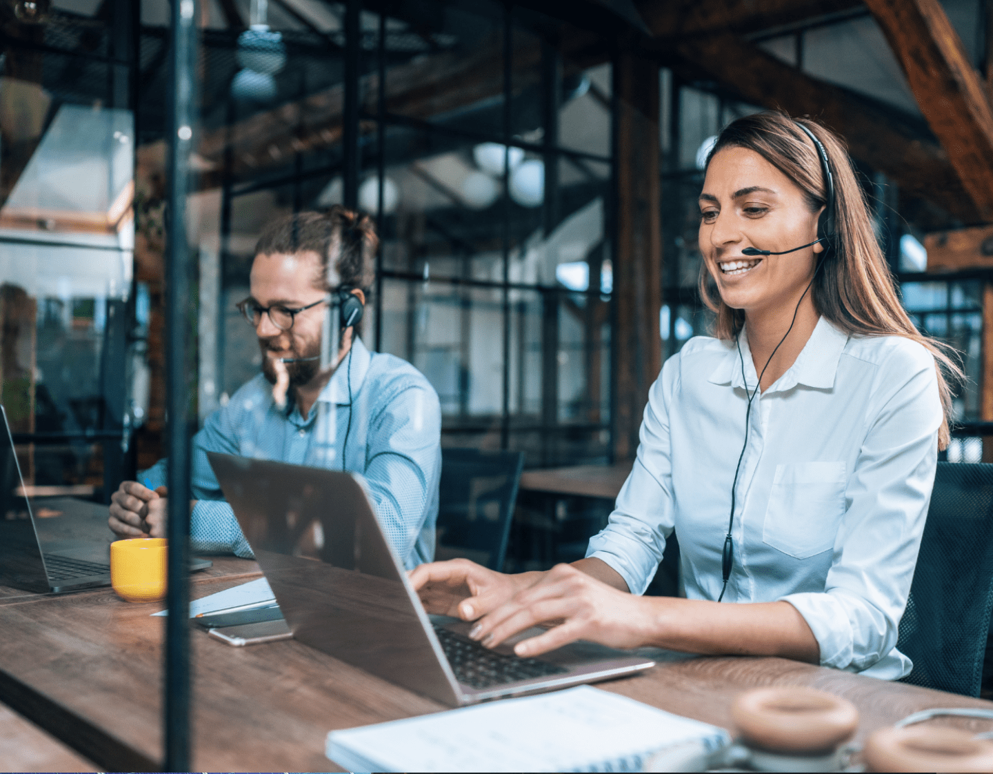 Two tech-support professionals speaking on headsets to help people with IT problems. They are sat at a wooden desk in a modern office.