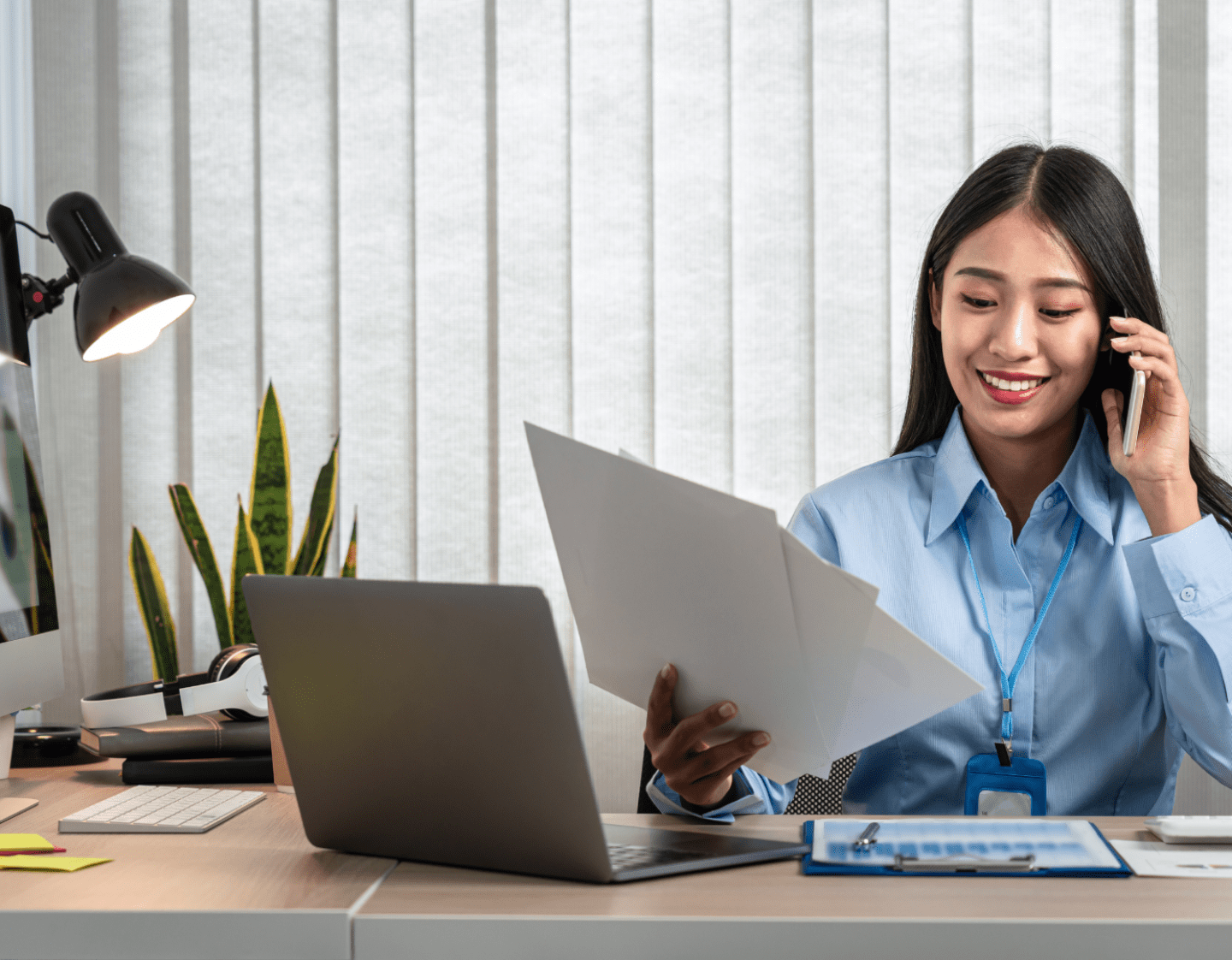 A woman sat at a desk making a phone call. She has her laptop open in front of her, ad 3 pieces of paper in her hand. 