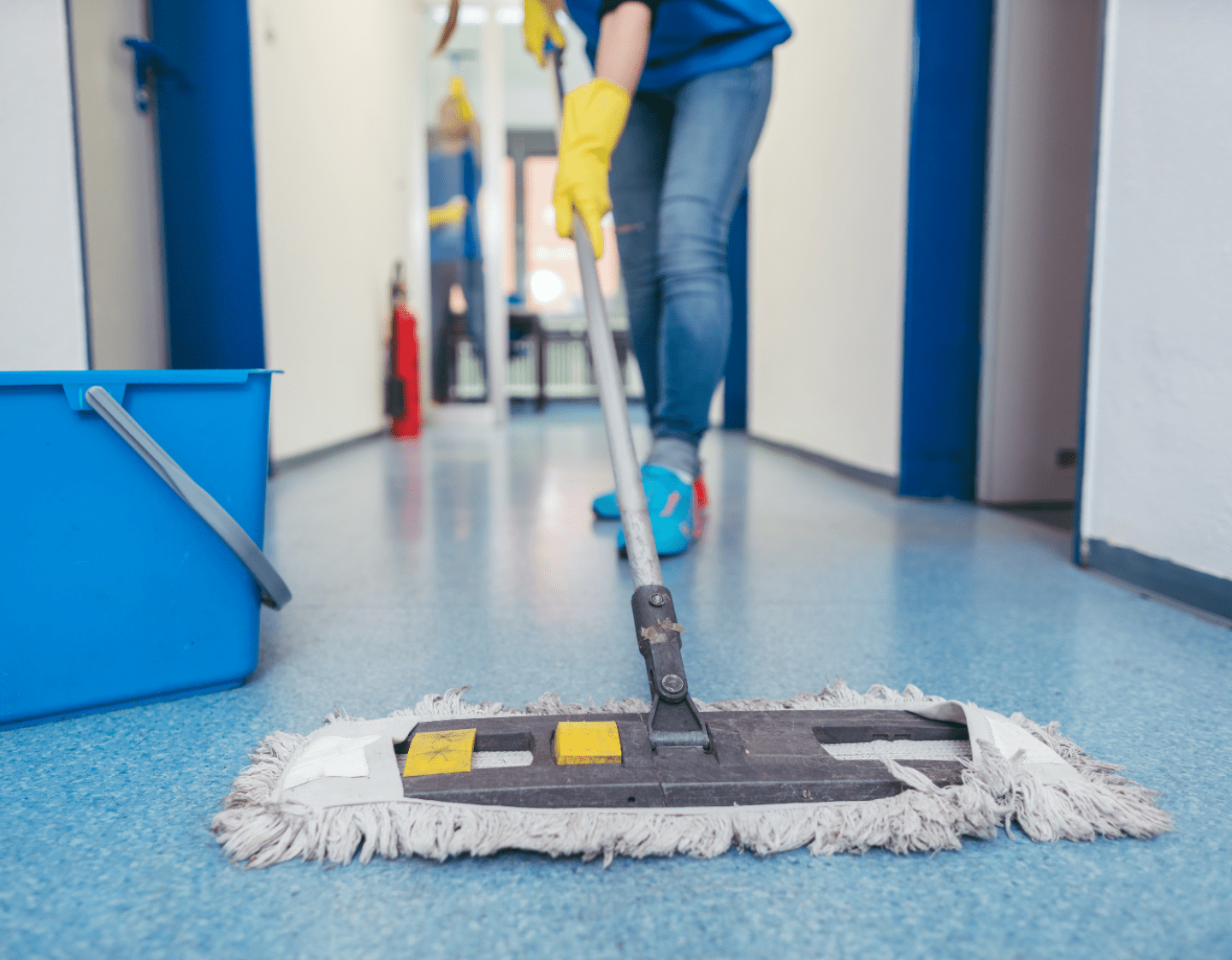 A close up image of a mop-head cleaning a floor. There is a bucket of water next to mop head, and a person in the background is holding the mop arm