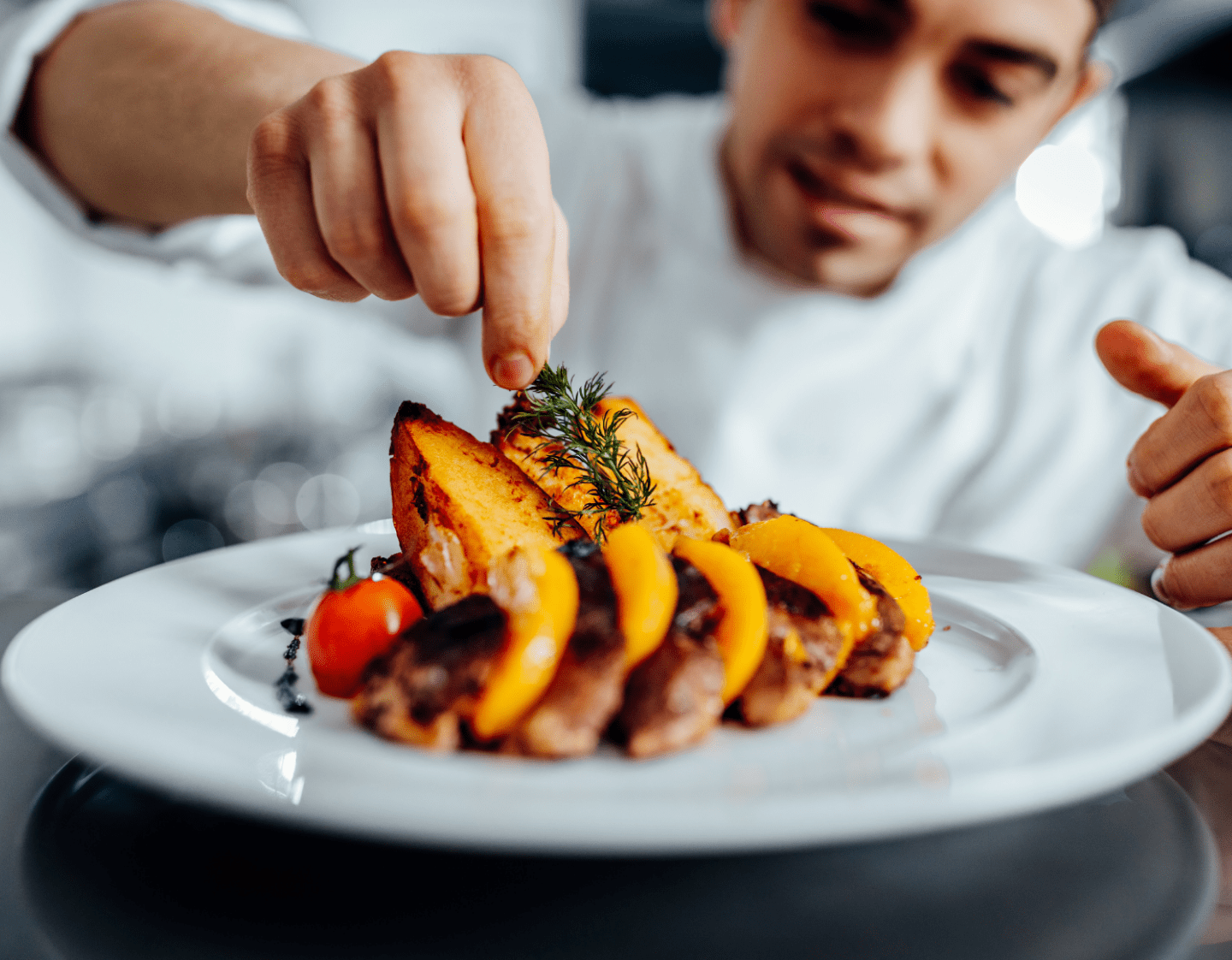 A chef adding garnish to a dish. The dish consists of potatoes, meat, tomatoes, and fruit.