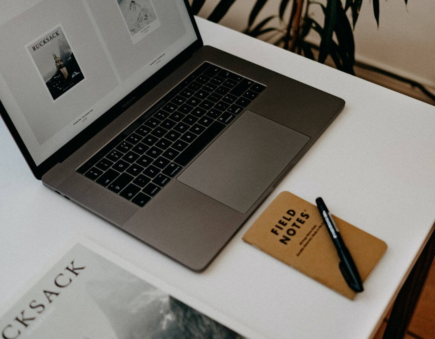 A laptop on a white desk with a brown notepad and pen next to it. The laptop screen shows an article being worked on