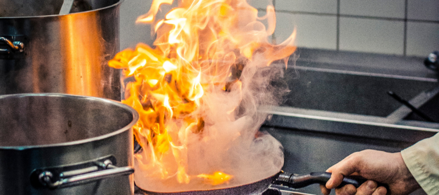 a frying pan on a stove top in a restaurant kitchen. Large flames are coming off the pan